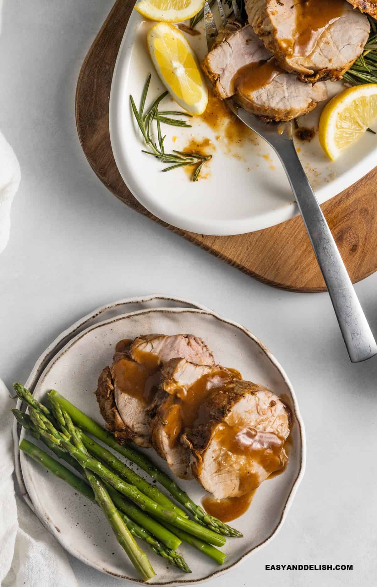 Sliced pork tenderloin with wine sauce served with asparagus and a platter in the background.