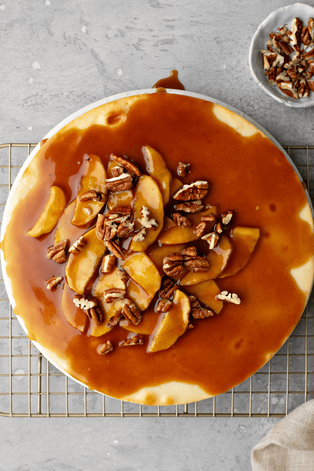 a whole caramel apple cheesecake on a rack with a bowl of pecans on the side.