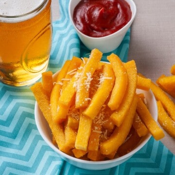 A bowl of polenta fries topped with Parmesan cheese with a side of ketchup and a drink.