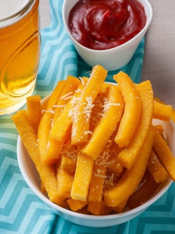 A bowl of polenta fries topped with Parmesan cheese with a side of ketchup and a drink.