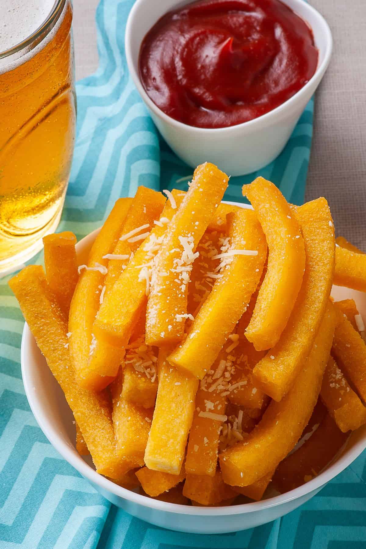 A bowl full of fried polenta fries with a glass of beer and a bowl of ketchup on the background. 
