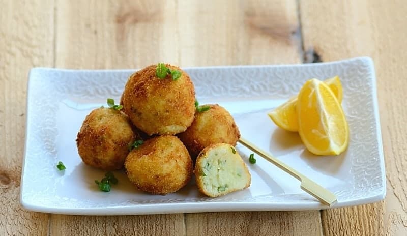 A stack of fried, golden-brown breaded codfish balls on a plate with a garnish of lemon slices 