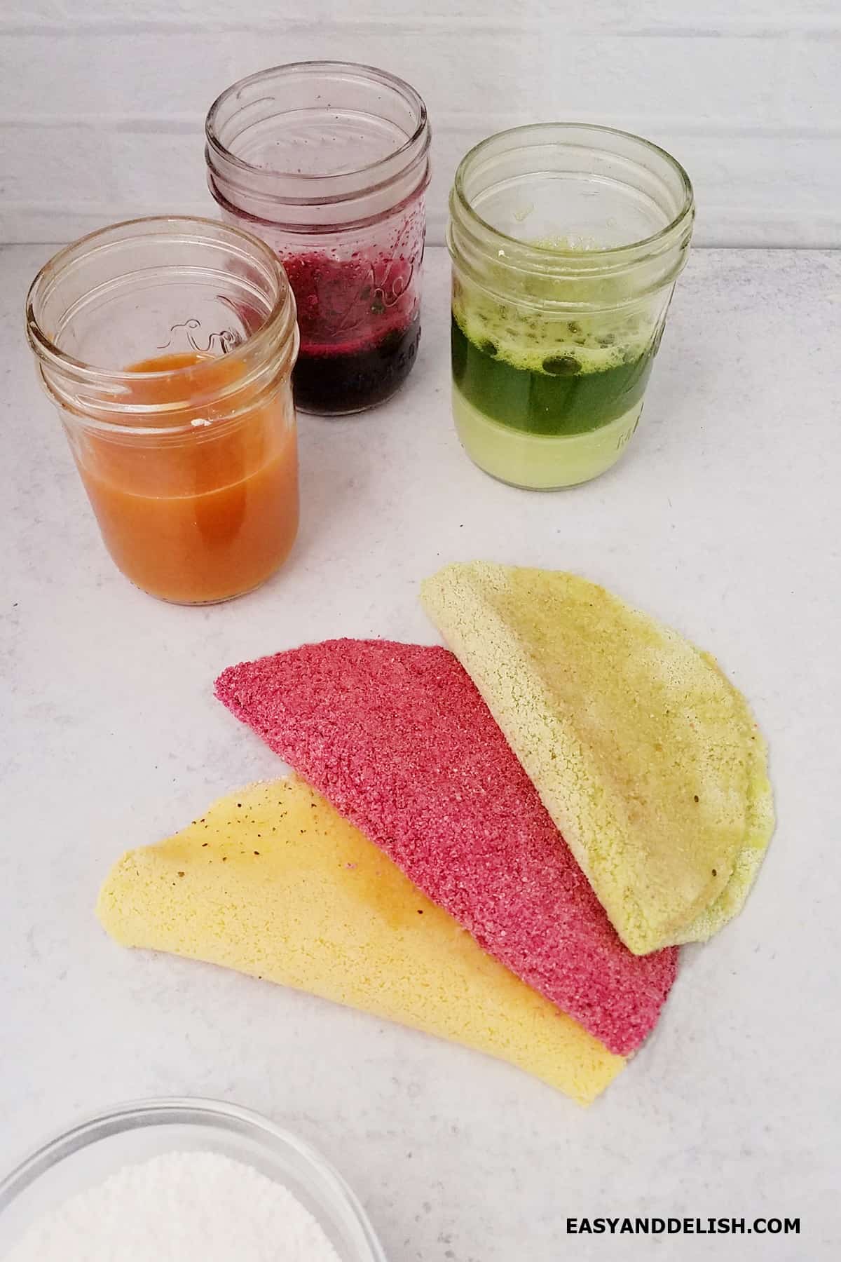 Three flavors of vegetable juices in jars and prepared Brazilian tapioca crepes nearby on a surface.