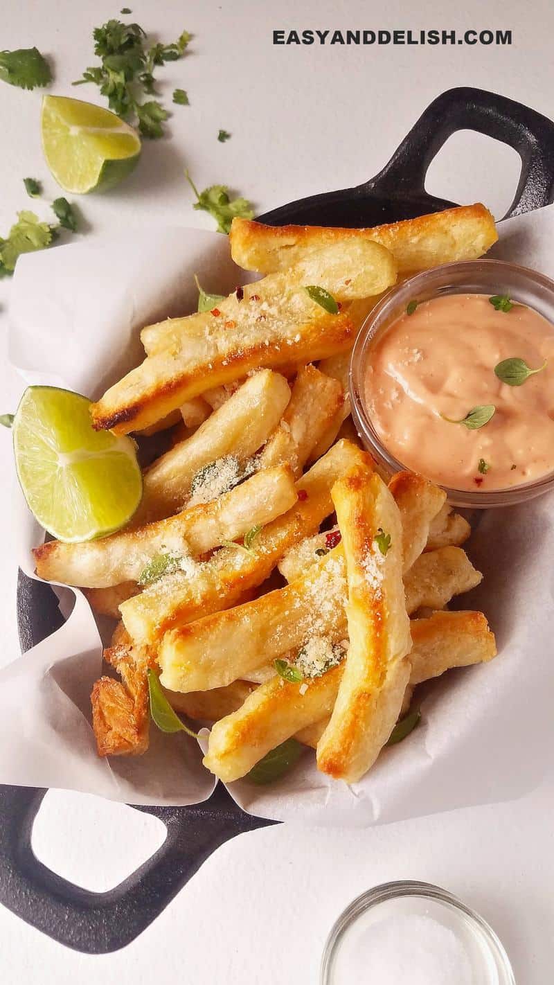 a bowl of yuca fries with a side of sauce and lemon wedges