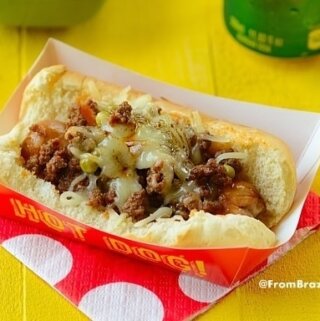 A deluxe hot dog on a bun topped with ground beef, cheese, and other toppings in a red paper tray