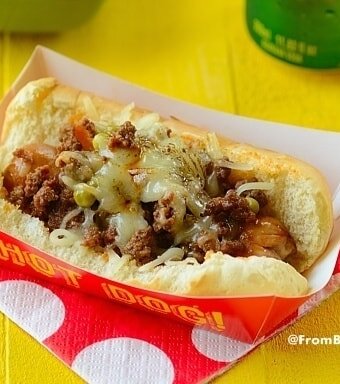A deluxe hot dog on a bun topped with ground beef, cheese, and other toppings in a red paper tray