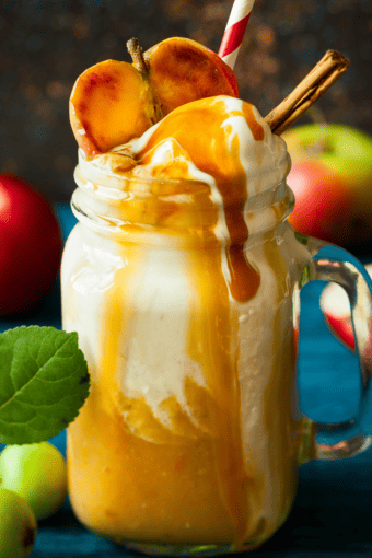 caramel apple cider in a tall glass topped with whipped cream and a dripping caramel sauce plus half an apple and cinnamon sticks.