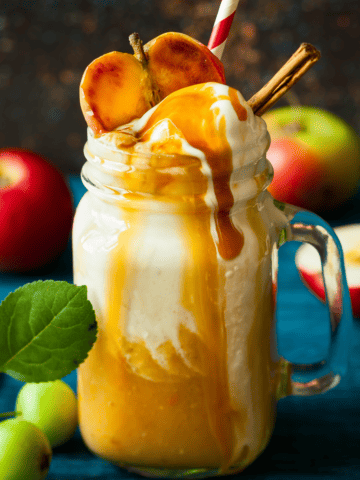 caramel apple cider in a tall glass topped with whipped cream and a dripping caramel sauce plus half an apple and cinnamon sticks.