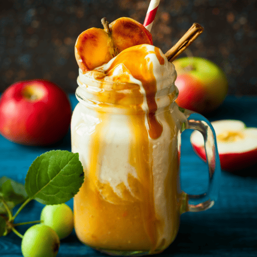 caramel apple cider in a tall glass topped with whipped cream and a dripping caramel sauce plus half an apple and cinnamon sticks.