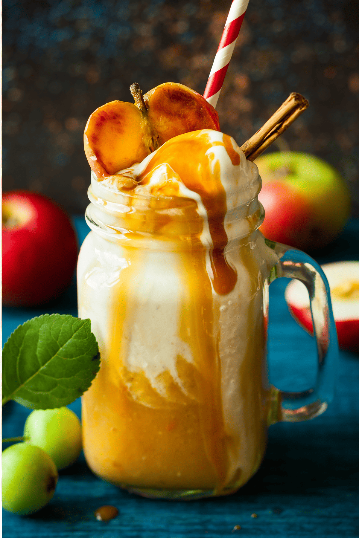 a glass of caramel apple cider topped with whipped cream and a dripping caramel sauce plus an apple, surrounded by apples and other garnishes.
