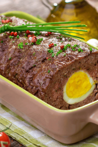 close up of German meatloaf with hard-boiled egg in a baking dish topped with red pepper and chives.