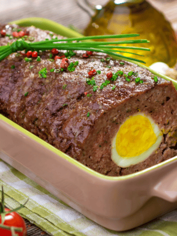 close up of German meatloaf with hard-boiled egg in a baking dish topped with red pepper and chives.