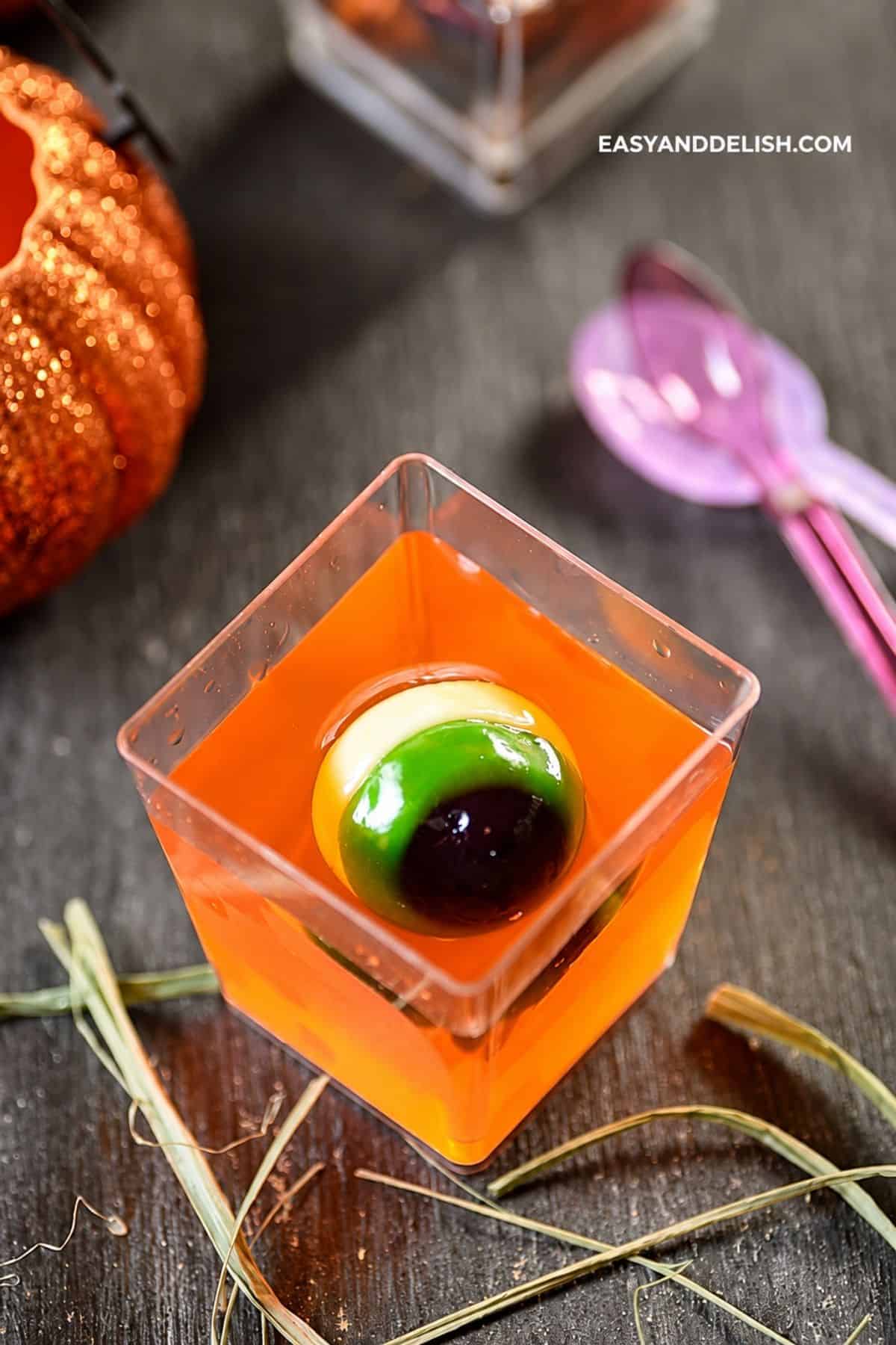 Creepy orange gelatin with a gummy eyeball in a clear square cup.
