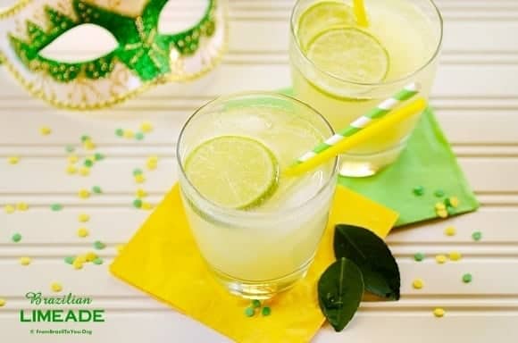 A glass of Brazilian limeade with ice cubes, garnished with a slice of lime, with a yellow straw and a green striped straw