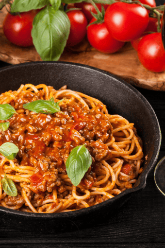 a skillet with spaghetti and meat sauce with tomato and fresh herbs on the background.