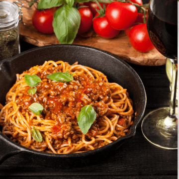 a skillet with spaghetti and meat sauce with tomato and fresh herbs on the background.