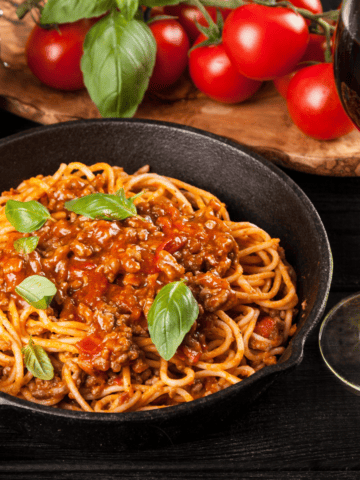 a skillet with spaghetti and meat sauce with tomato and fresh herbs on the background.