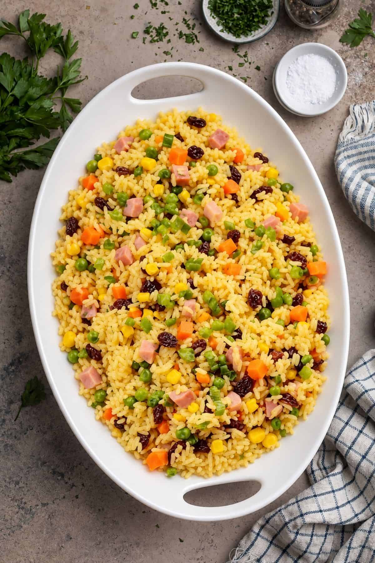 Yellow rice mixed with diced ham, carrots, peas, corn, green onions, and raisins on a white oval platter.