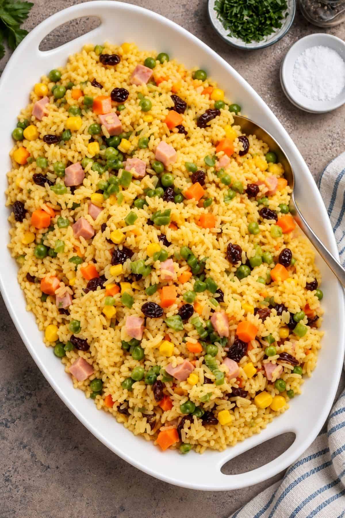 Close-up of yellow rice with diced ham, peas, carrots, corn, and raisins beside a metal serving spoon.