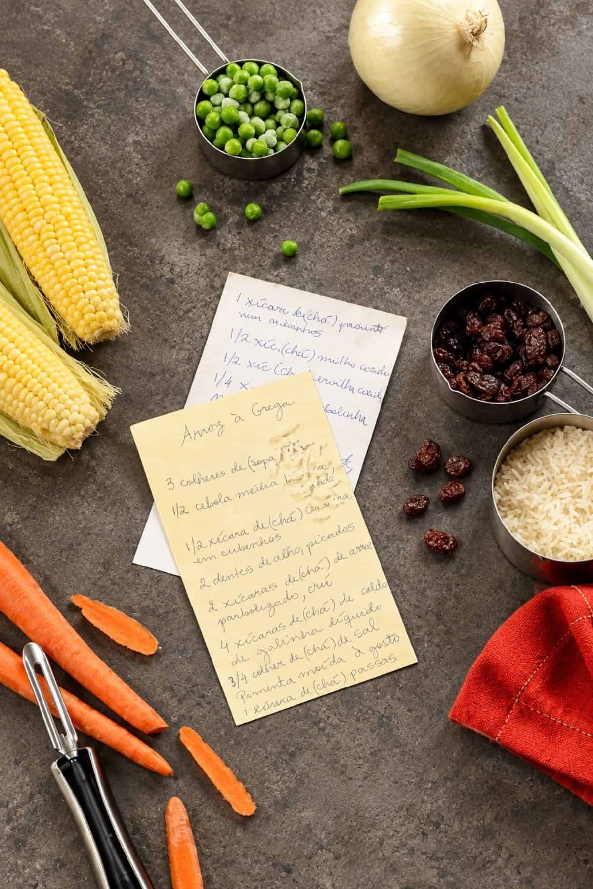 Handwritten Brazilian Arroz à Grega recipe cards surrounded by corn, peas, onion, raisins, rice, and carrots.