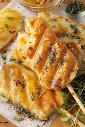 CLOSE UP OF SEVERAL GRILLED CHEESE ON A STICK SERVED ON A LARGE TRAY WITH RED PEPPER FLAKES, HONEY AND FRESH HERBS.