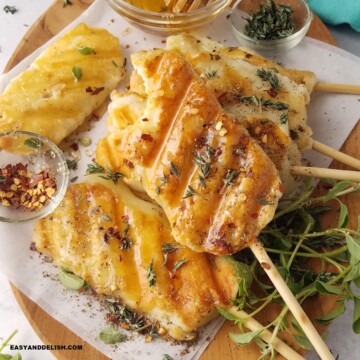 CLOSE UP OF SEVERAL GRILLED CHEESE ON A STICK SERVED ON A LARGE TRAY WITH RED PEPPER FLAKES, HONEY AND FRESH HERBS.