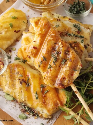 CLOSE UP OF SEVERAL GRILLED CHEESE ON A STICK SERVED ON A LARGE TRAY WITH RED PEPPER FLAKES, HONEY AND FRESH HERBS.