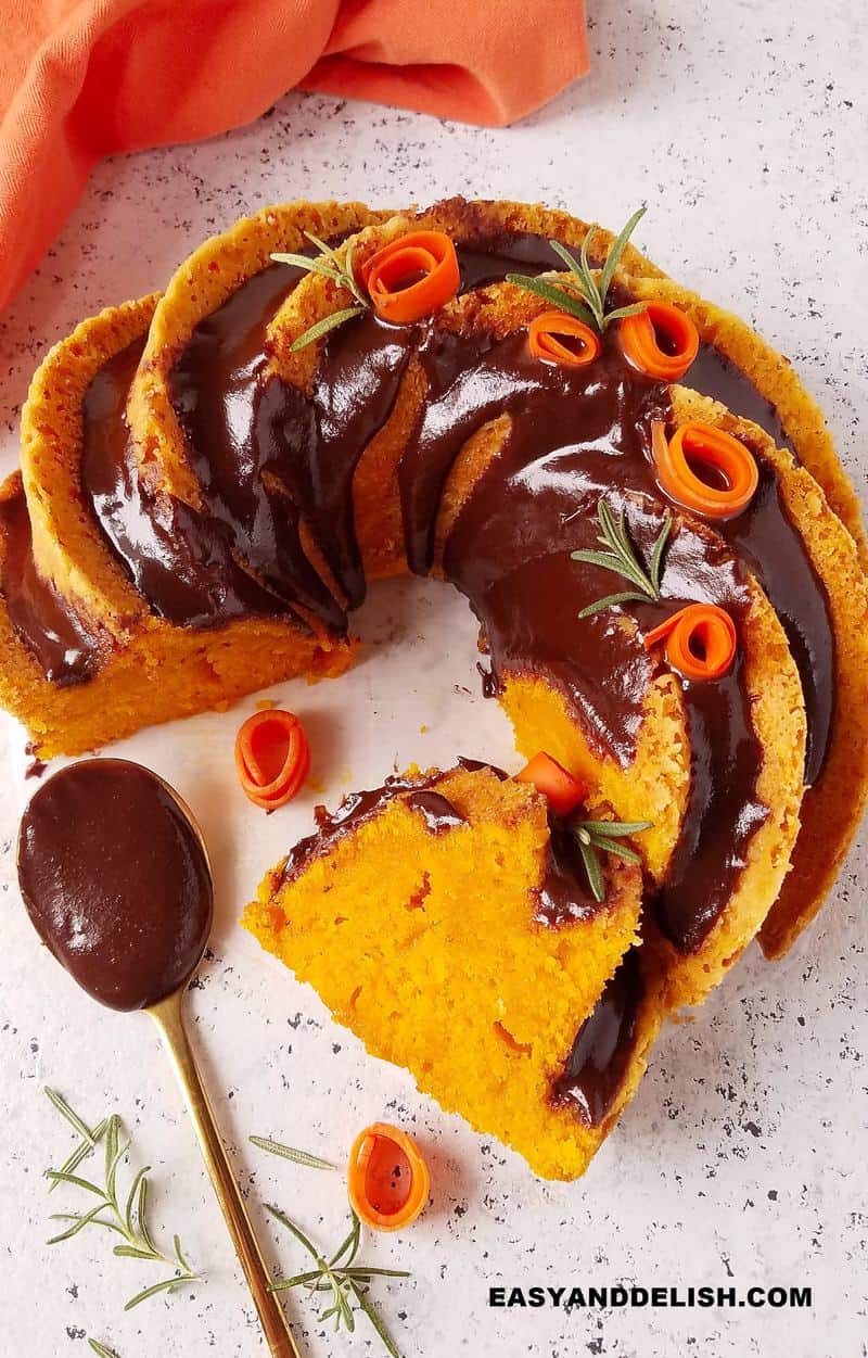 bolo de cenoura fofinho parcialmente fatiado com uma colher de cobertura de chocolate ao lado