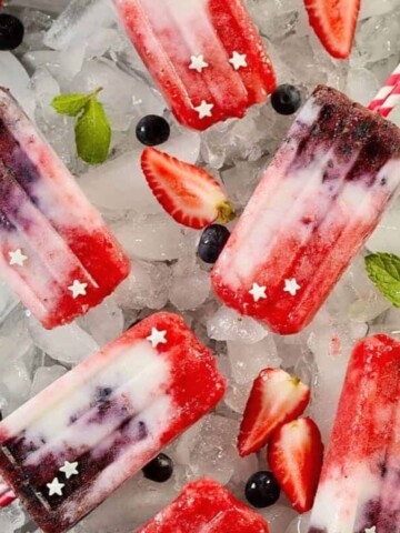 Several red, white, and blue berry yogurt ice pops on ice cubes with some scattered ingredients around them.