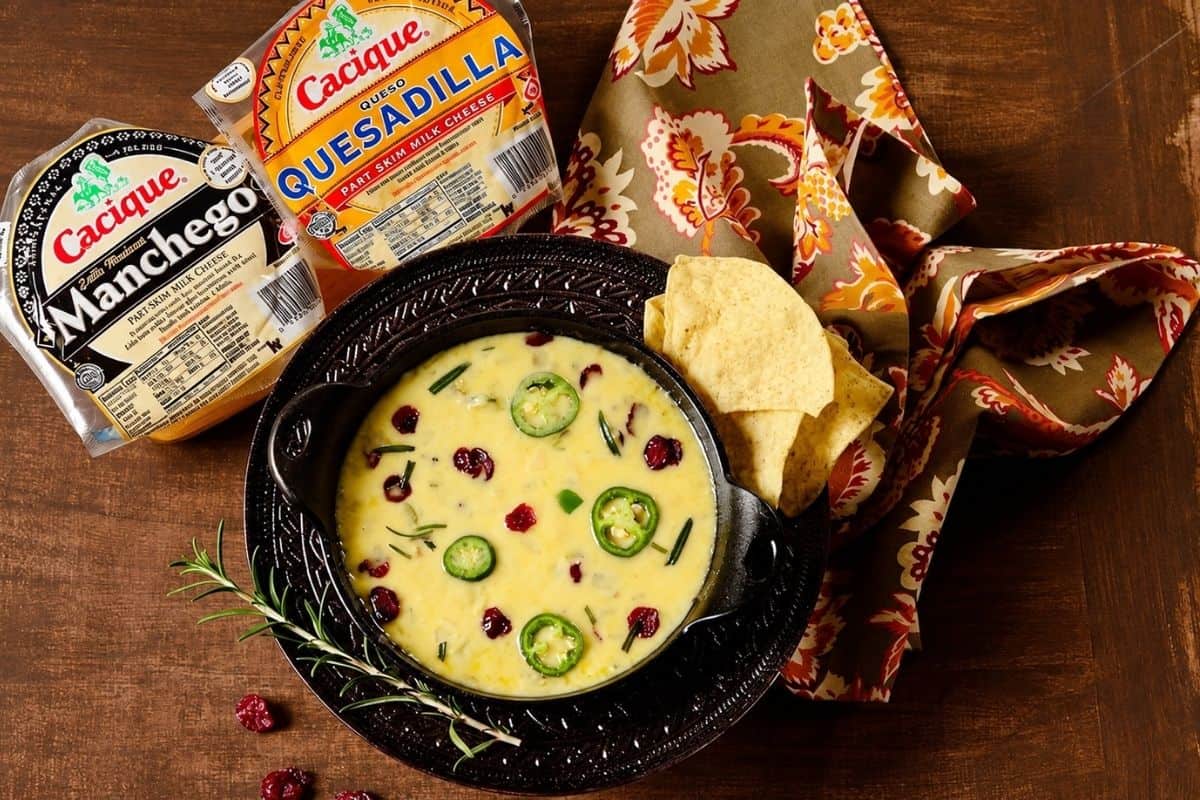 Queso fundido dip in a black skillet with tortilla chips, rosemary, dried cranberries, jalapeños, and Manchego and queso quesadilla packages.