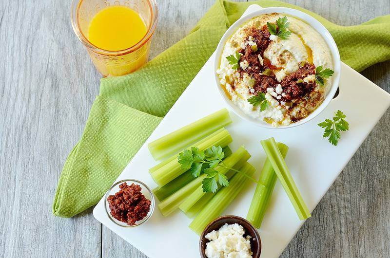 veggies and a bowl of dip topped with crumbled cheese and chorizo plus a glass of juice.