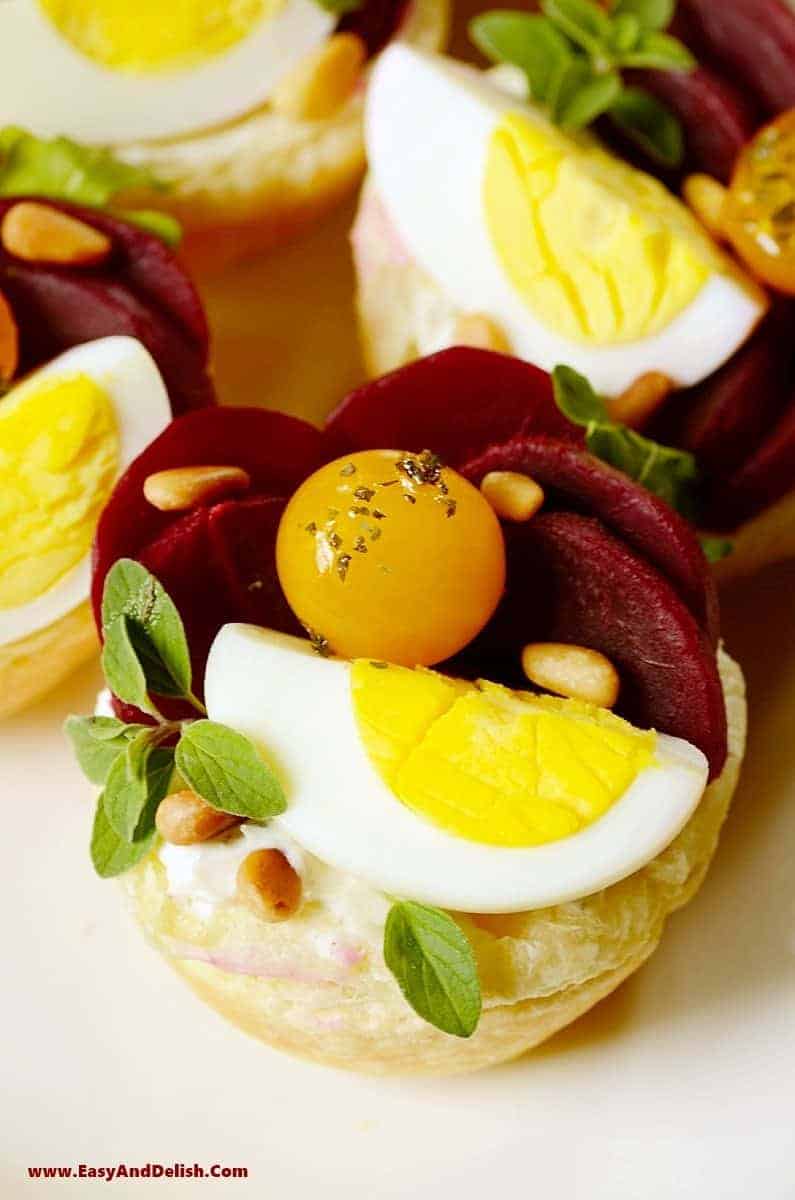 a close up of one puffy pastry tartlet topped with boiled eggs, beet slices, pine nuts, and cherry tomatoes