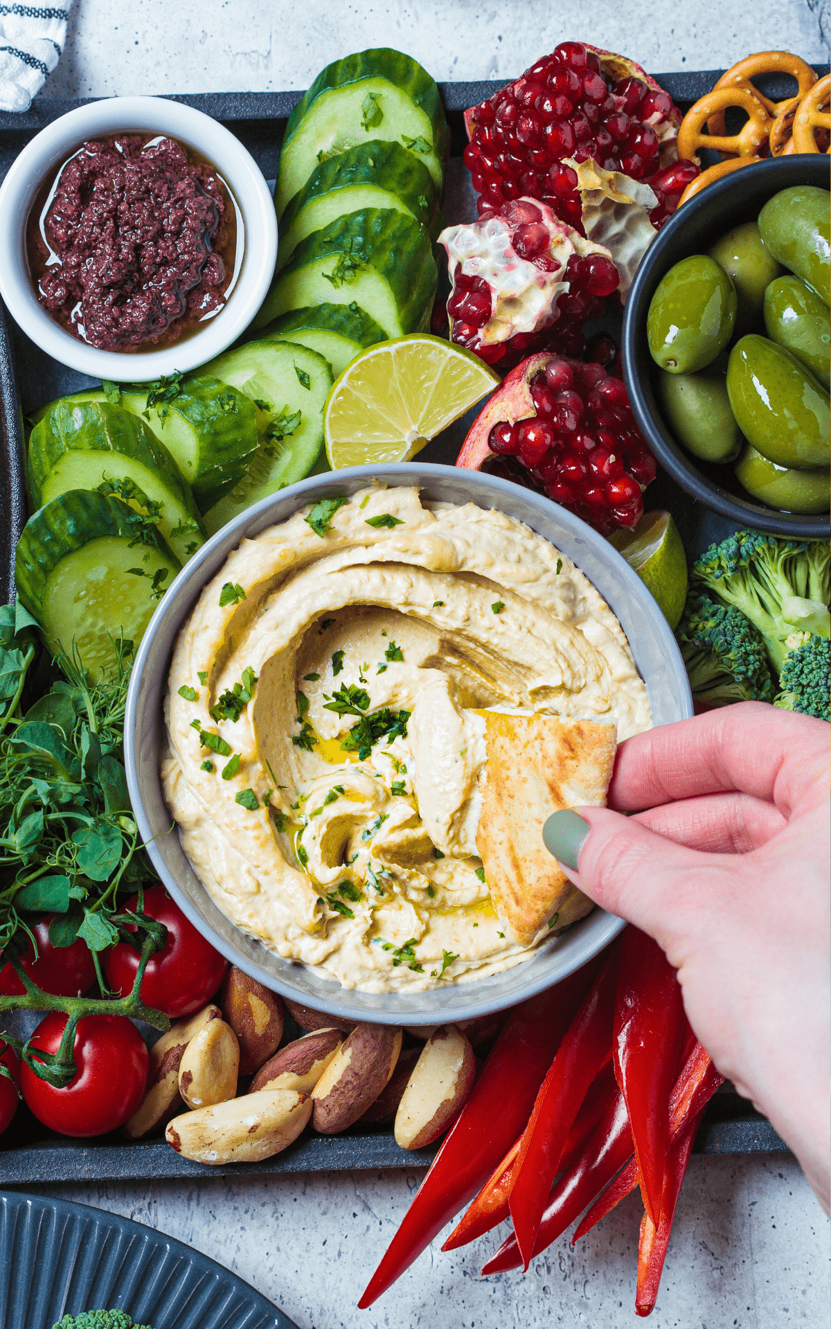 close up of a bowl of keto hummus with a hand dipping pita chips in it and veggies around it.