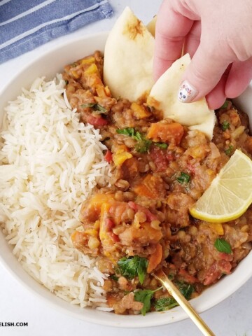 Close up of a bowl of Instant Pot lentils, Moroccan style, with vegetables and warm spices. A hand dips a piece of naan in the lentil stew.