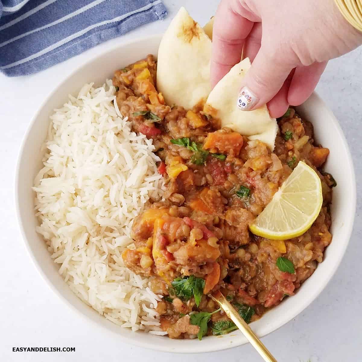 Close up of a bowl of Instant Pot lentils, Moroccan style, with vegetables and warm spices. A hand dips a piece of naan in the lentil stew.