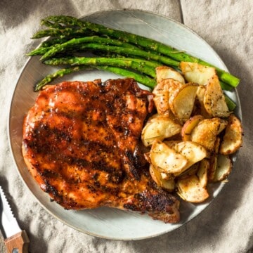A Blackstone griddled pork chop in a plate with a side of asparagus and crispy potatoes.