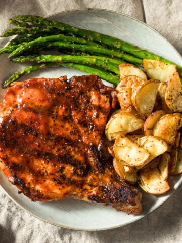 A Blackstone griddled pork chop in a plate with a side of asparagus and crispy potatoes.