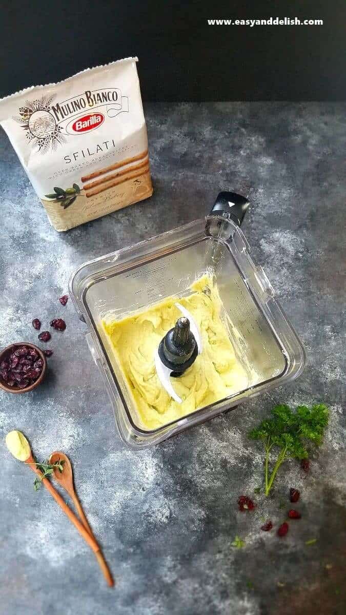 Avocado Hummus Recipe is a no cooking, quick and easy appetizer made with a few simple ingredients, and served with olive breadsticks. It’s perfect for the Holidays! avocado hummus being blended in a blender with breadstick package on the background