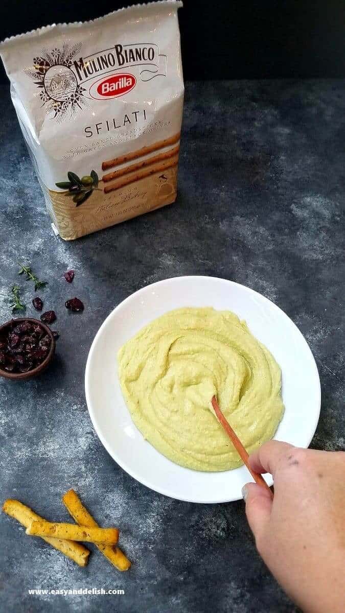 Avocado Hummus Recipe is a no cooking, quick and easy appetizer made with a few simple ingredients, and served with olive breadsticks. It’s perfect for the Holidays! mixing avocado hummus dip with a spoon and breadsticks on the side