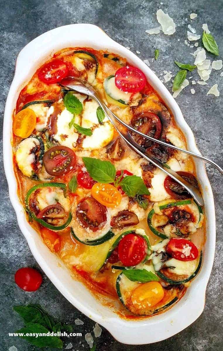 Overhead view of a baking dish containing baked Caprese zucchini roll ups with silverware on the side