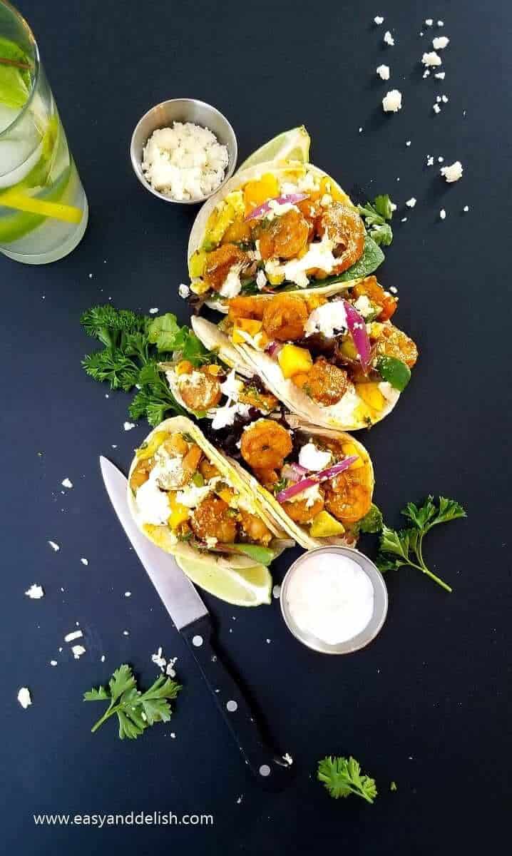 Overhead view of Mojito shrimp street tacos lined up between bowls of garnishes, a knife, and a drink glass. 