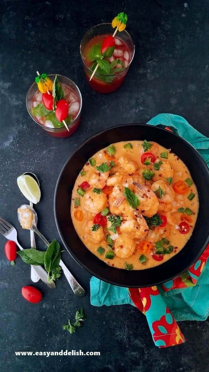 Overhead view of a bowl of Brazilian shrimp stew (ensopado de camarao) with silverware and 2 glasses of a drink near the bowl 