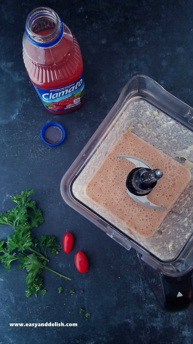 Image showing vegetables blended in a blender with additional vegetables and a bottle of Clamato juice near the blender. 