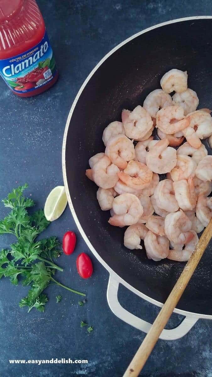 Image of a pan containing sauteed shrimp -- with lime, vegetables, and Clamato juice near the pan. 