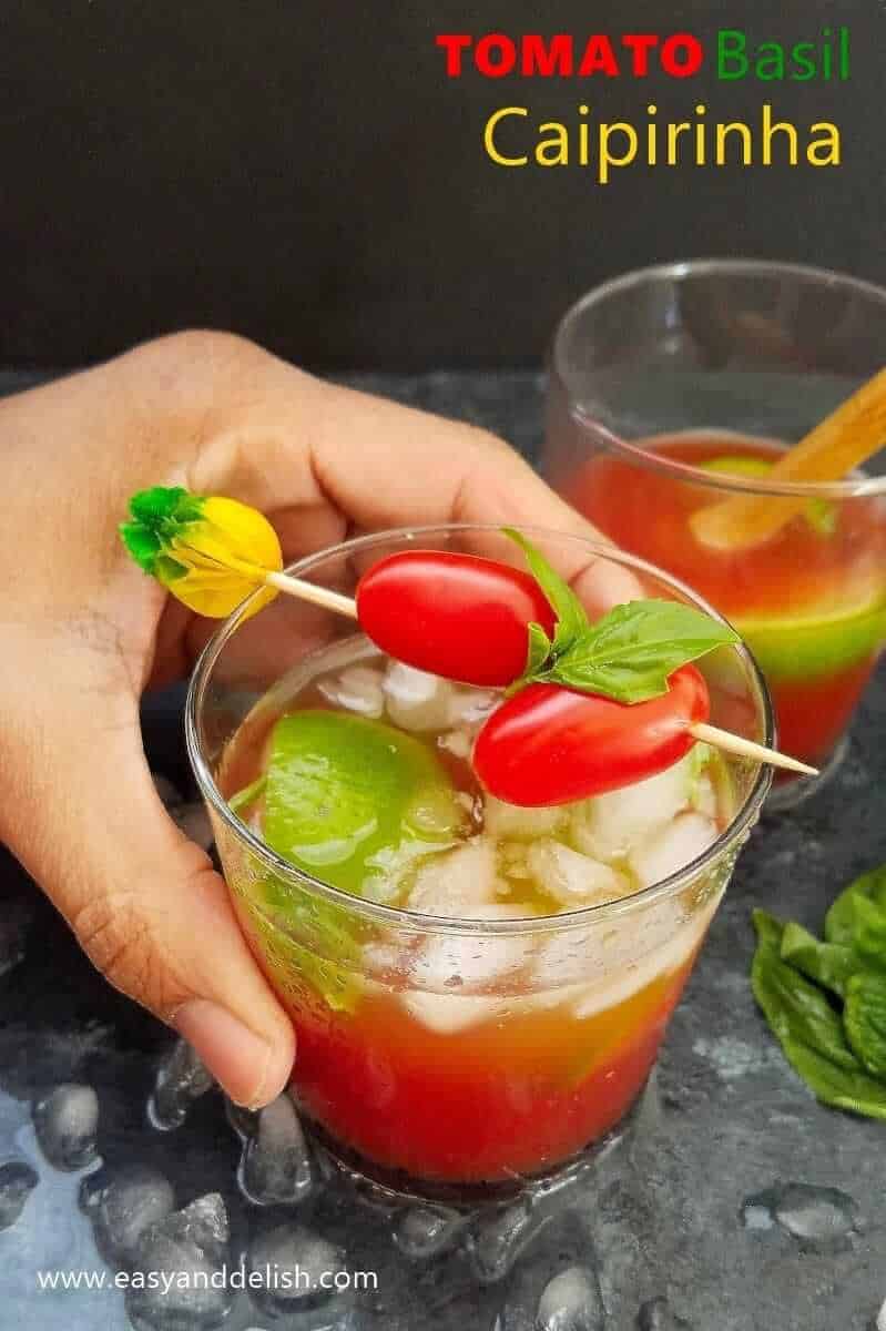 Image of 2 glasses filled with tomato basil caipirinha with a hand holding one of the glasses. 