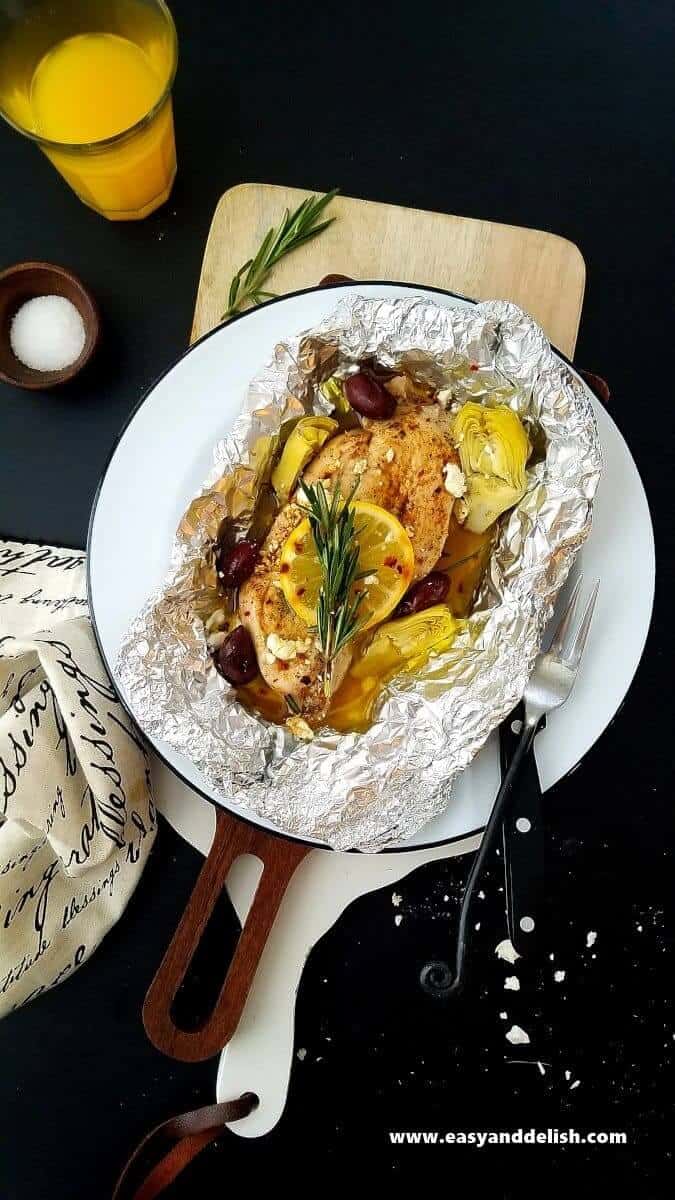 Overhead view of Mediterranean Chicken Foil Packets over a plave with silverware on the side served with a glass of juice and garnishing