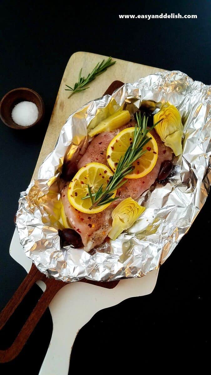 Image of a Mediterranean Chicken Foil Packet opened, showing the layering process (lemon slices and vegetables) right before baking in the oven. 