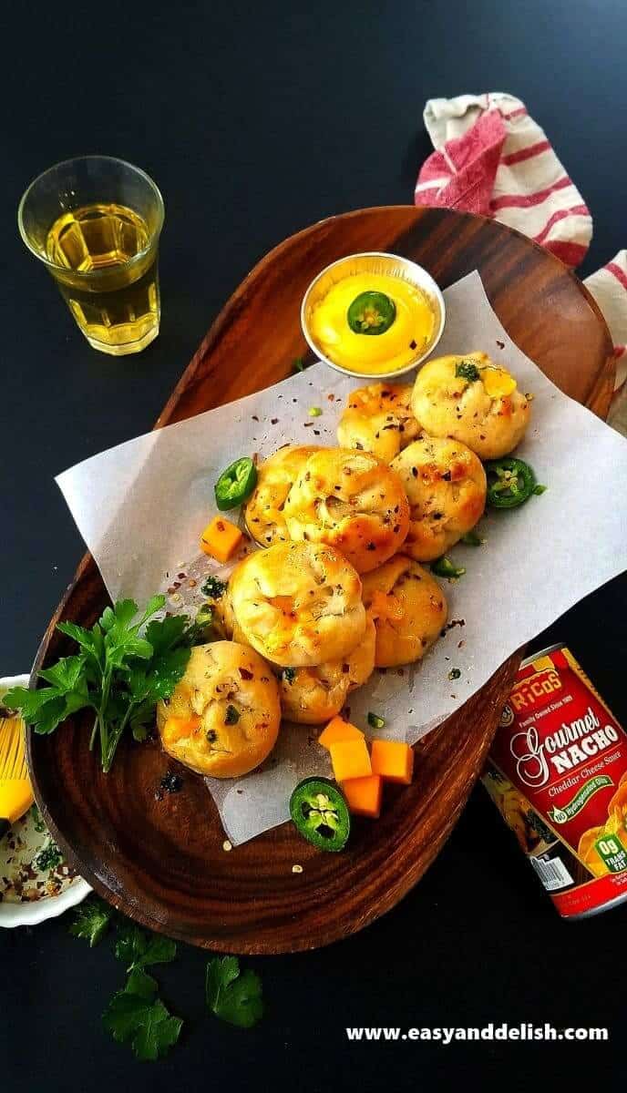 Image of nacho pizza bombs on a platter with a small bowl of nacho sauce. A can of nacho sauce and a glass of beer are shown near the platter. 