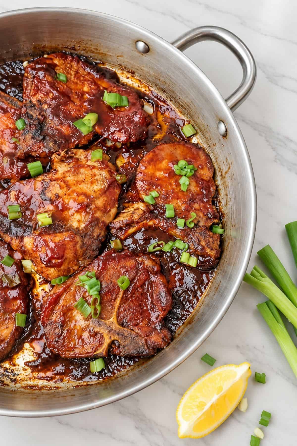 Barbecue Pork Chops coated in glossy reddish-brown sauce in a stainless steel skillet, garnished with chopped green onions.