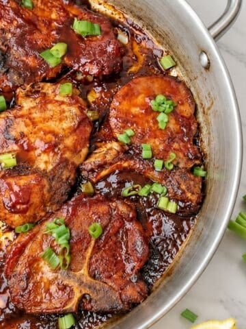 Pork chops coated in barbecue sauce with chopped green onions in a stainless steel skillet.